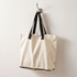 A beige foldable tote bag with black straps hanging on a hook. It features a logo.