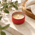 A red mini tin candle sits on a stone surface, with a flickering flame surrounded by greenery and a wooden tray.
