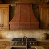 Custom copper kitchen hood in a rustic kitchen with warm patina, distressed wood cabinets, and stone countertops