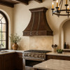 Decorative copper kitchen vent hood with silver vertical straps in Spanish Revival inspired kitchen, hammered copper canopy above arched openings and warm stone accents