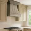 Natural zinc range hood with curved bell canopy in bright transitional kitchen with shaker cabinets, stone backsplash and large window