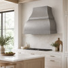 Hammered zinc bell range hood in a bright minimalist kitchen with pale wood, stone backsplash, and large window