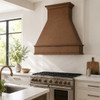 Hammered copper range hood in a modern kitchen with marble surfaces, white cabinetry, and large window