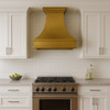 Brass range hood with flared silhouette mounted above a stainless steel stove in a transitional kitchen with white shaker cabinets and subway tile backsplash