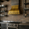 Brass trapezoid range hood with riveted straps in an industrial kitchen with dark cabinets and brick walls