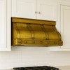 Undermount brass range hood with riveted straps and trapezoid silhouette in modern kitchen, planished brass finish creating warm contrast with sleek cabinetry
