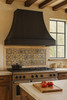 Black zinc kitchen range hood with trapezoidal canopy in a Spanish revival kitchen with tile backsplash and wood ceiling beams