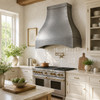 Bright French country kitchen with a hammered zinc bell range hood, cream cabinetry, brass, and a window