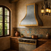 Zinc range hood with brass corner plates and straps in a modern farmhouse kitchen with painted cabinets and stone counter