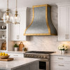 Zinc range hood with decorative brass corners and straps in a rustic European kitchen with marble and muted cabinetry