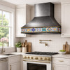 Gunmetal range hood with Talavera tile apron above stove in bright suburban kitchen with white cabinets and window