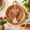 Handmade Floral Mexican Heart Ornament with Pink Tassel displayed on a decorative woven wall basket