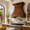 Hammered copper range hood with flared crown and curved apron in a Mediterranean kitchen with blue tile backsplash