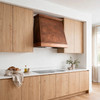 Sloping copper range hood with stepped crown in a Scandinavian warm minimalist kitchen with light oak cabinetry, soft neutral finishes, and natural stone countertops
