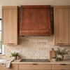 Sloping copper range hood with stepped crown in a contemporary rustic kitchen with reclaimed wood cabinetry, natural stone countertops, and warm copper tones