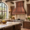 Bell copper range hood with forged iron straps in Spanish Colonial kitchen with white plaster, dark beams, and tile