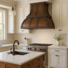 Wall mount copper range hood for gas stoves in transitional kitchen, hammered bell shaped canopy with decorative straps above balanced cabinetry and refined stone surfaces