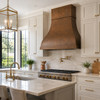 Hammered copper range hood in a bright transitional kitchen with white Shaker cabinets, quartzite, brass hardware and tall windows
