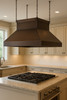 Chain suspended hammered copper island range hood in Victorian style above kitchen island with gas stove, tapered canopy with decorative chains and warm patina finish