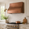 Smooth box copper range hood in transitional kitchen with neutral cabinets, stone backsplash, and mixed metal accents