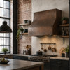 Hammered box copper range hood in industrial loft kitchen with exposed brick, black cabinetry, and steel shelving