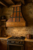 Copper range hood with dark straps in rustic kitchen, riveted bands over warm copper patina above range and stone backsplash