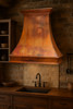 Country kitchen copper exhaust hood in rustic kitchen, curved copper canopy with warm rustic patina above wood beams and stone backsplash
