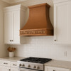 Hammered copper exhaust hood in a transitional kitchen with mixed metals, neutral stone, and integrated grapevine apron