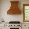 Wall-mount hammered copper exhaust hood in a modern farmhouse kitchen with Shaker cabinets and grapevine apron