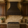 Farmhouse copper range hood with decorative scroll apron in Mediterranean villa style kitchen, hammered bell canopy with warm antique patina above stone surfaces