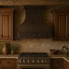 Farmhouse copper range hood with decorative scroll apron in rustic kitchen, hammered bell canopy with warm antique patina above wood beams and stone backsplash