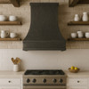 Dark zinc range hood with riveted vertical strap seams in a rustic contemporary kitchen with warm wood, textured stone, and rich artisan patina