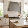 Hammered zinc range hood in a transitional kitchen with white Shaker cabinets, marble, and brass hardware