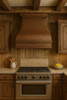 Copper range hood with apron scroll in rustic kitchen, smooth canopy with decorative band above range and wood beams and stone backsplash