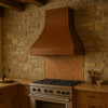Copper range hood with smooth apron in rustic kitchen, hammered canopy with warm patina above range and wood beams and stone backsplash
