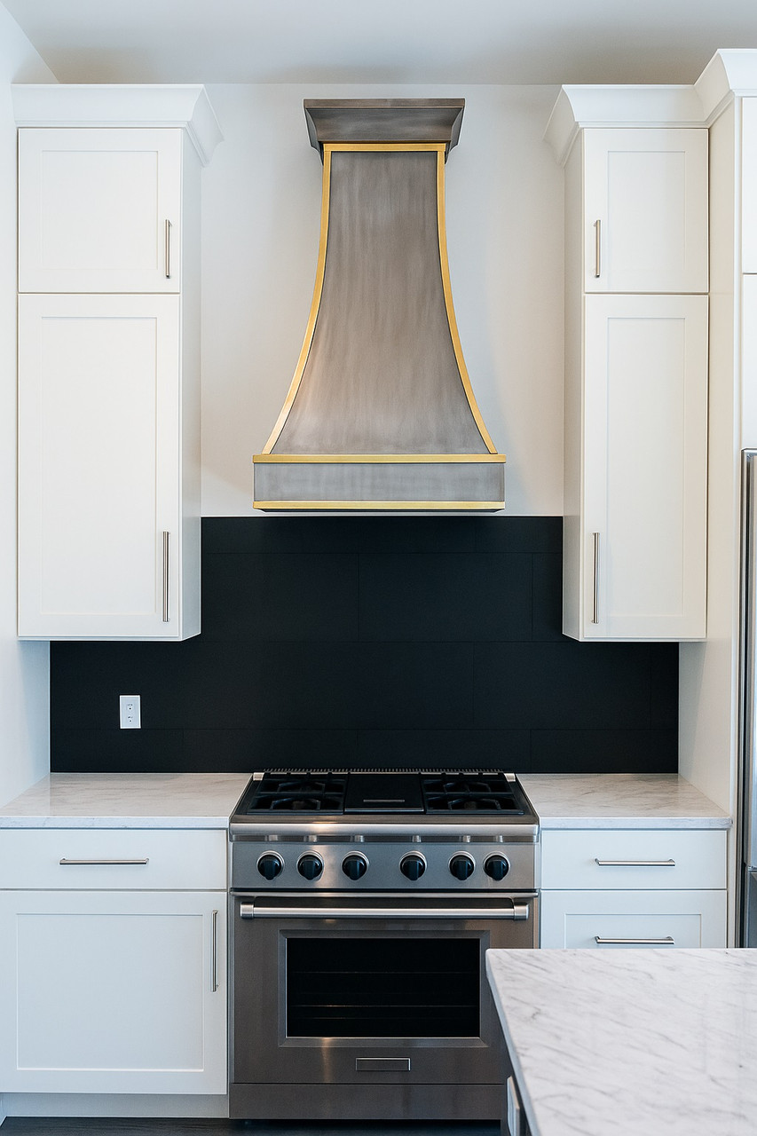 Brass range hood with golden accents in a contemporary style kitchen with minimalist white cabinets and black backsplash