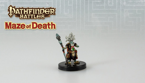 This pale blue humanoid has bright white eyes and wild, white hair adorned with a spiked, gem-encrusted coronet. Dressed in messy robes covered in ornate baubles, one hand holds a tall staff. (Front View)