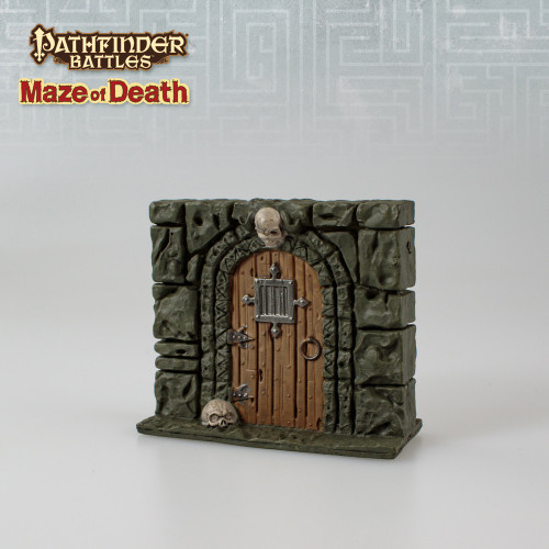 An image of a heavy wooden door set into a stone wall, a skull used as the doorframe's keystone. The top center of the door has a barred metal window. (Front View)