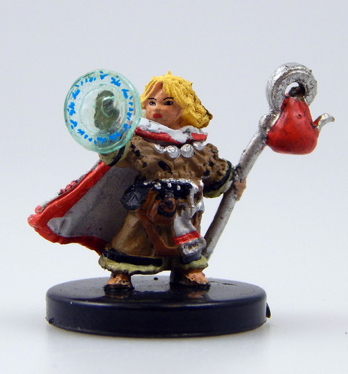 An image of a stoic blonde halfling dressed in several layers of furs and a thick white cloak lined in red. In her left hand she holds a silver staff topped with a hollow circle through which a red bindle is tied. Her right hand is held up in a ring of magical runes casting a spell. (Front View)