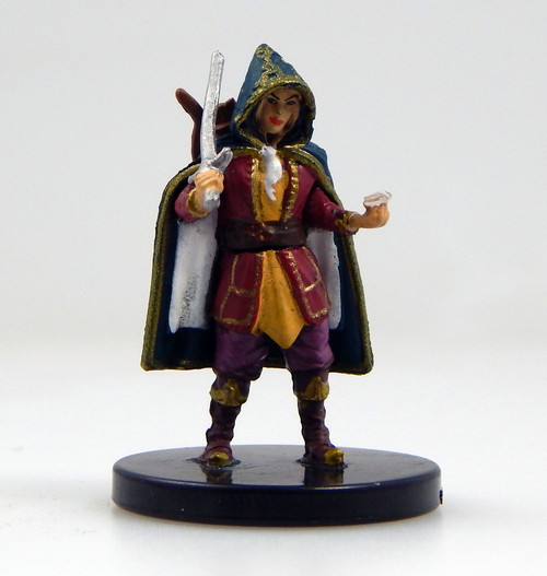 An image of a thief guildmaster in brightly colored, noble clothes and a muted blue, hooded cloak with gold embroidery. Smirking, in one hand she holds a large gemstone while she rests a sword on her shoulder with the other. (Front View)