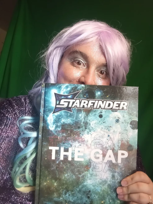 Starfinder: THE GAP photo shows a pink haired person holding the book in front of their face so they’re peeking over the top of the book; the book cover shows a swirling churn of mist and stars and planets and darkness.