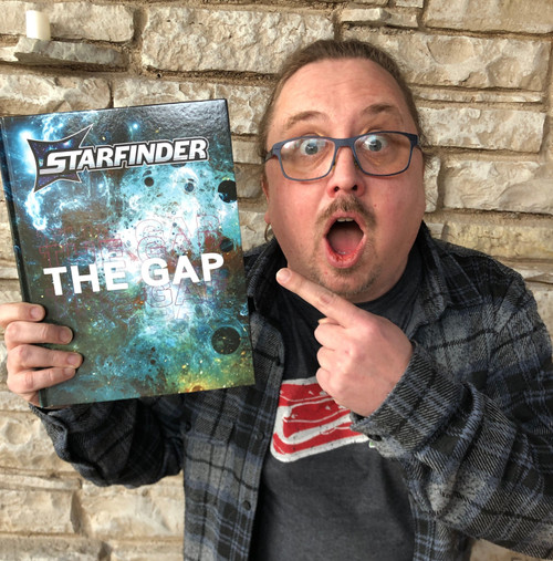Starfinder: THE GAP photo shows an incredulous man in a flannel shirt and glasses holding the book up and pointing at it while standing in front of a brick wall; the book cover shows a swirling churn of mist and stars and planets and darkness.