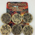 Pathfinder Hero Point Tokens (Pack of 6) shows a six brass-colored coins sitting atop a cardboard label featuring the Pathfinder Hero Point Tokens logo and an image of heroes fighting a dragon, but the image is mostly covered by the six coins; three coins show a stylized capital letter P, while the other three show a vaguely “X” shaped spiky symbol under a four-pointed star.