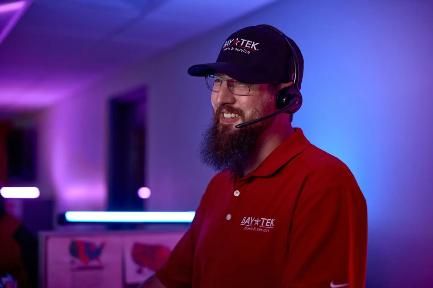 BayTek Employee with a beard wearing a headset, cap, and red polo shirt, smiling in an office with purple lighting.