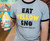 Close-up of a person wearing the gray 'EAT YELLOW SNO' t-shirt, holding a cup of yellow shaved ice.