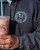 Close-up of a person holding a drink while wearing the gray zip-up hoodie, showing the small circular logo on the chest.