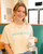 A smiling young woman with blonde hair wearing an off-white "Avalanche Crew" t-shirt, holding a Bahama Buck’s drink cup inside a shop.