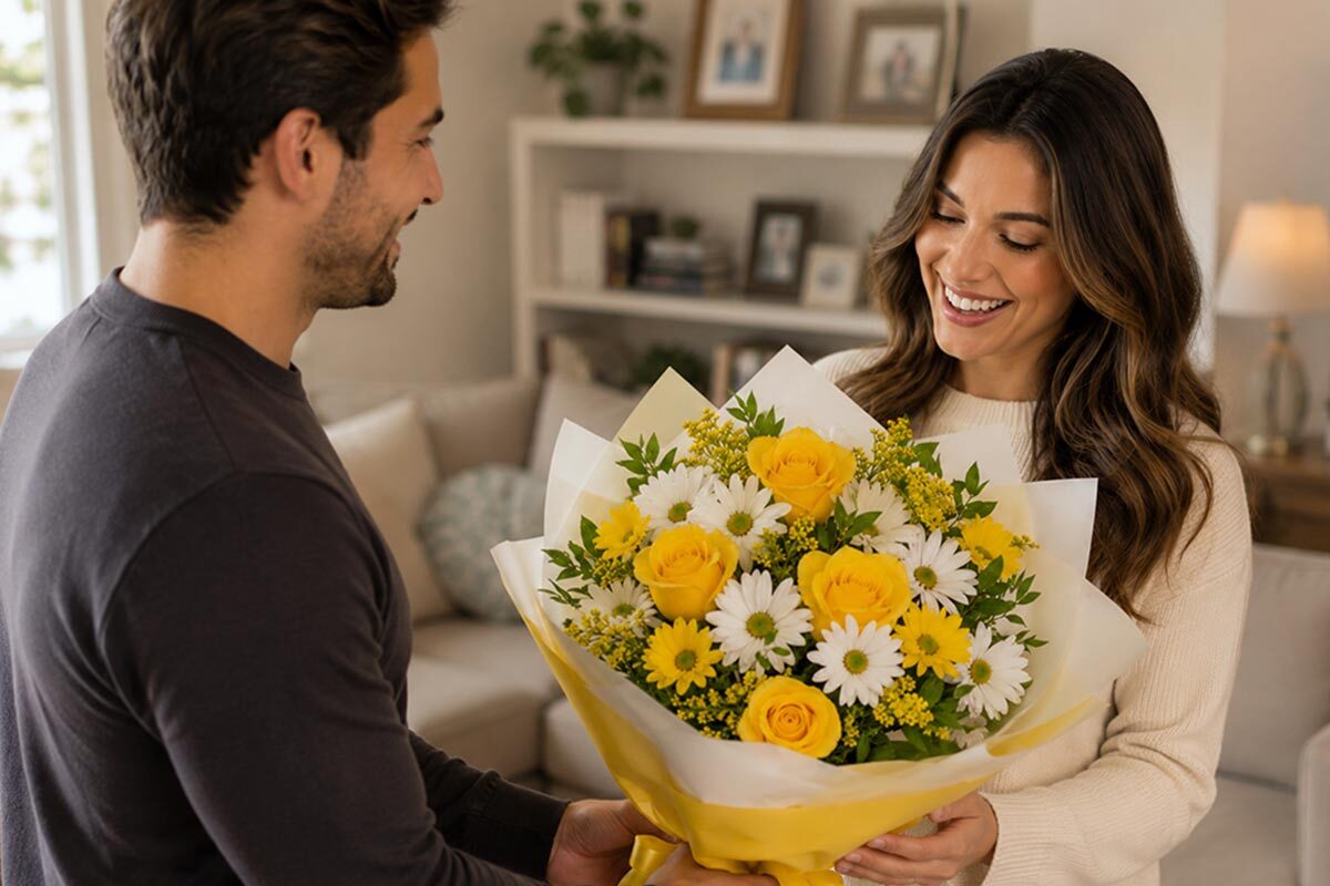 Husband giving a bright yellow and white flower bouquet wrapped in yellow paper to his smiling wife in a cozy living room.