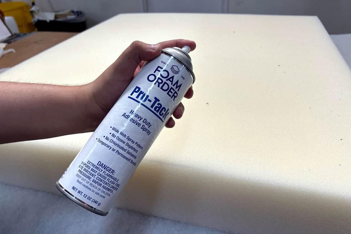 Pro-Tack Foam Adhesive, a can of heavy-duty adhesive spray with a white and blue label, being held over a large piece of foam material.