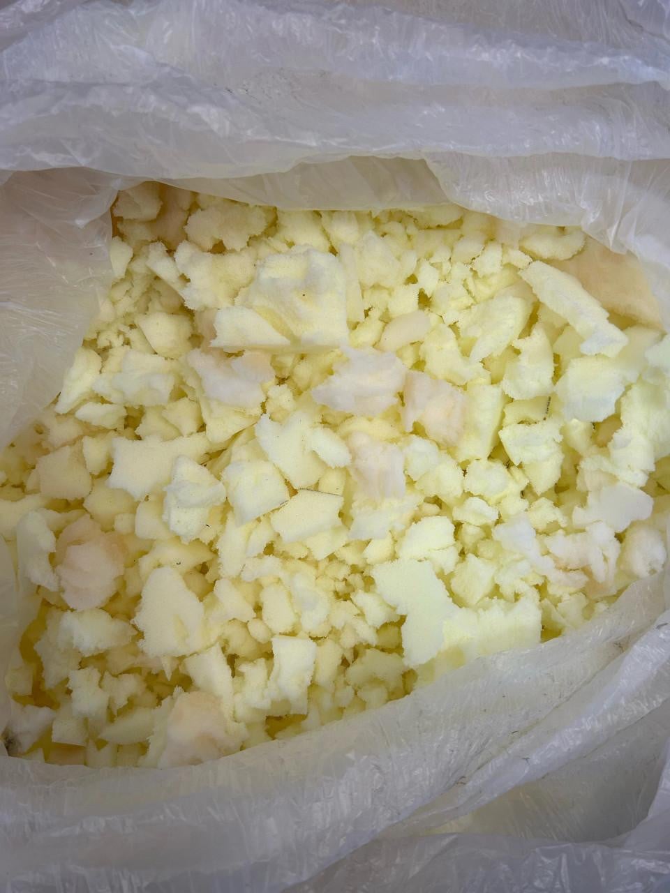 Shredded Polyurethane Foam, close-up view showing small, irregular pieces of soft, yellowish foam material inside a transparent plastic bag.